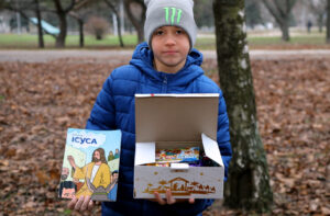 Ukrainian boy in a park holding an open Gift of Hope Ukraine box filled with sweets and a colorful Christian Scripture book.
