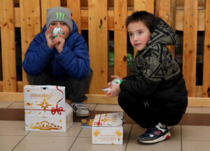 Two Ukrainian boys crouched on a tiled floor, opening and enjoying small treats next to their decorated Gift of Hope boxes.
