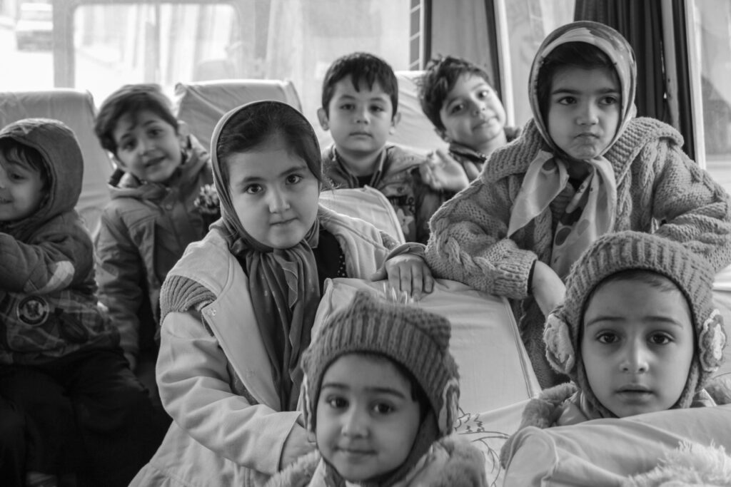 A group of young children wearing winter hats and coats, looking toward the camera in a crowded indoor setting.