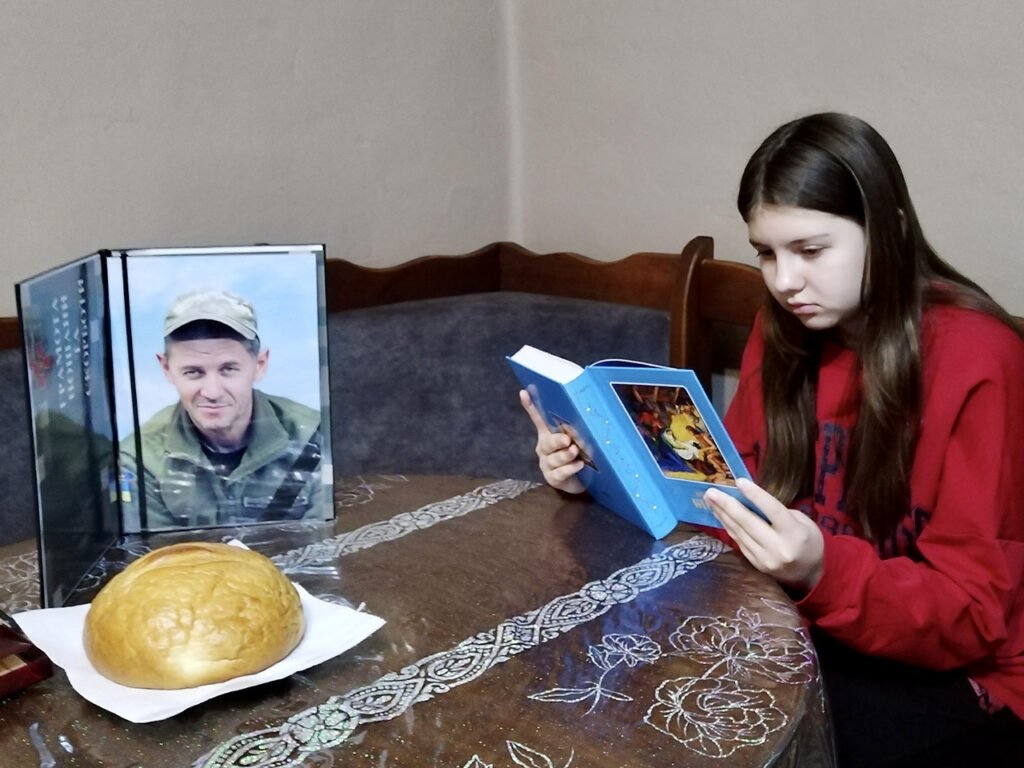 A young girl in a red sweater sitting at a table, looking down sadly at a framed photograph of a man in a military uniform and a loaf of bread.