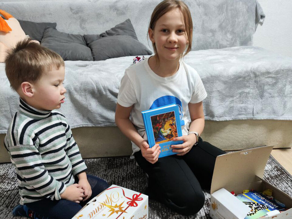 Two young children sitting on the floor indoors, with a girl holding a colorful book and a young boy looking at an open gift box containing various items.