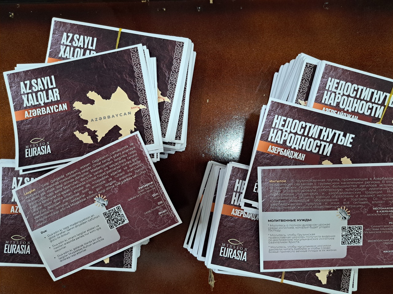 Stacks of Mission Eurasia prayer cards for unreached people groups in Azerbaijan, featuring maps, cultural information, and QR codes for ministry support.