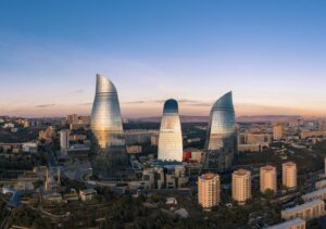 A wide aerial view of the Baku city skyline at sunset, featuring the three modern Flame Towers overlooking the Caspian Sea and smaller residential buildings.