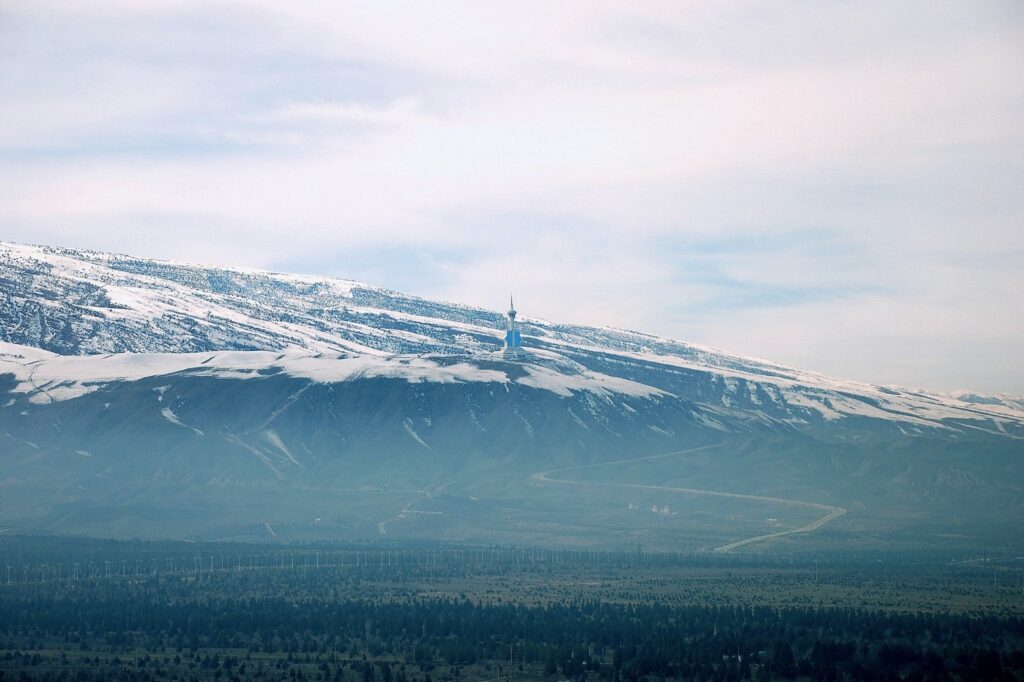 A wide scenic view of the snow-capped Kopet Dag mountains under a hazy sky, overlooking a green valley with a winding road and a distant monument.
