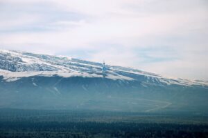 A wide scenic view of the snow-capped Kopet Dag mountains under a hazy sky, overlooking a green valley with a winding road and a distant monument.