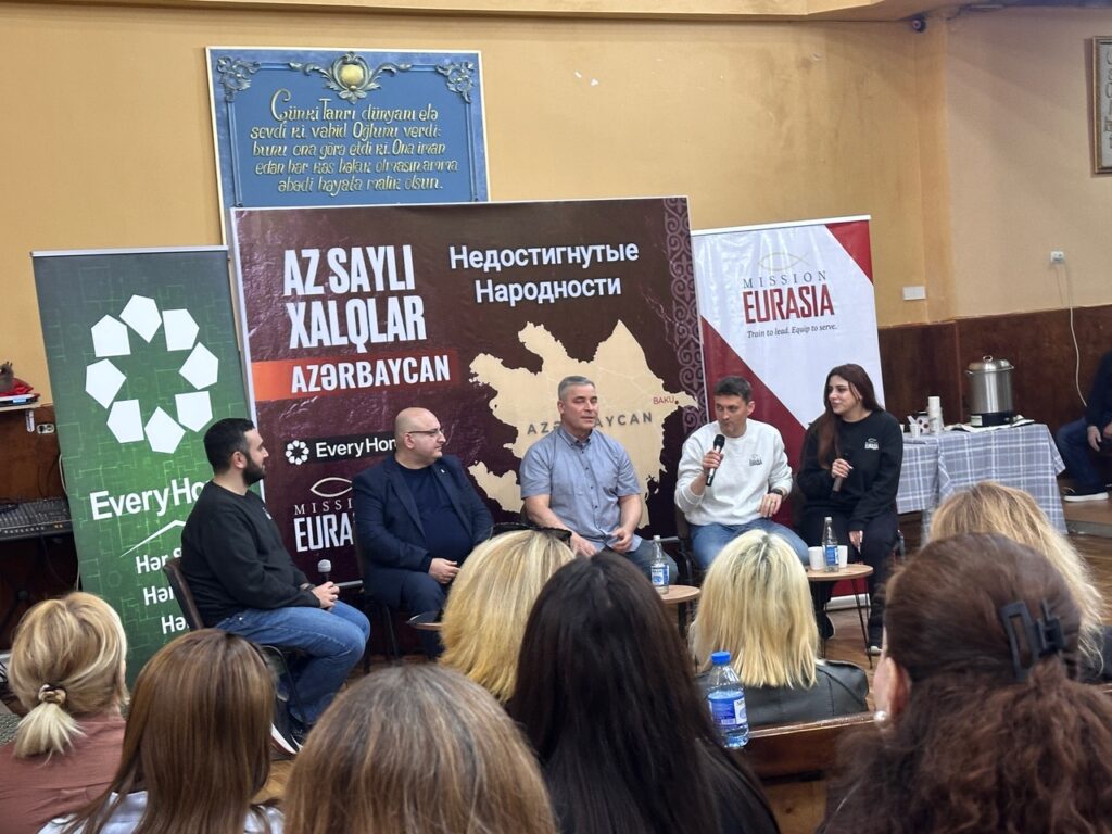 A panel of ministry leaders speaking at a Mission Eurasia event in Azerbaijan, with banners displaying maps of unreached people groups and ministry branding in the background.