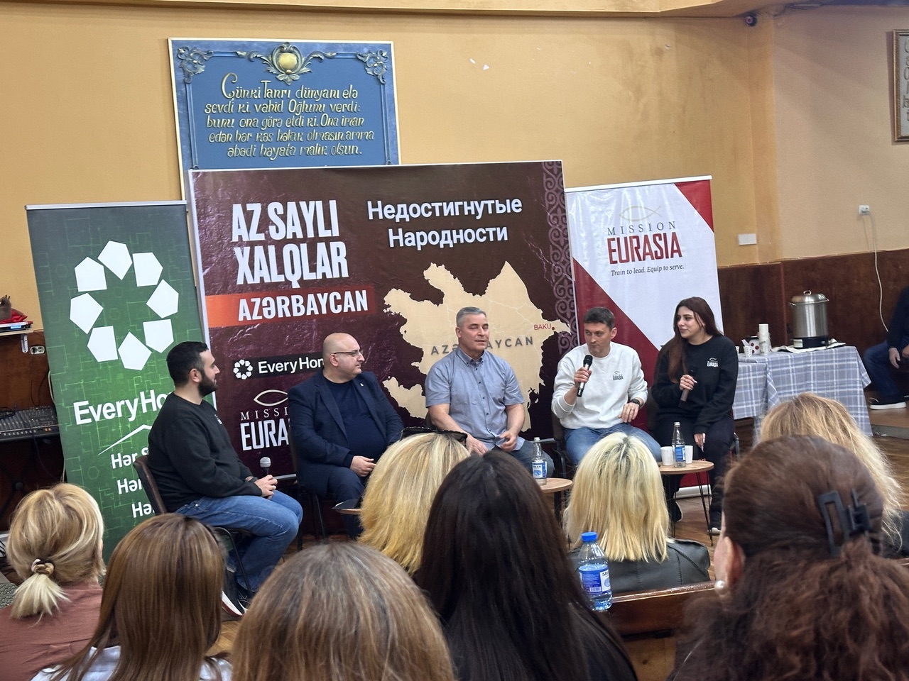 A panel of ministry leaders speaking at a Mission Eurasia event in Azerbaijan, with banners displaying maps of unreached people groups and ministry branding in the background.