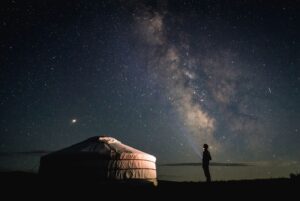 A traditional Mongolian ger (yurt) illuminated from within at night, sitting under a vast, star-filled sky with the Milky Way visible and a person standing nearby.