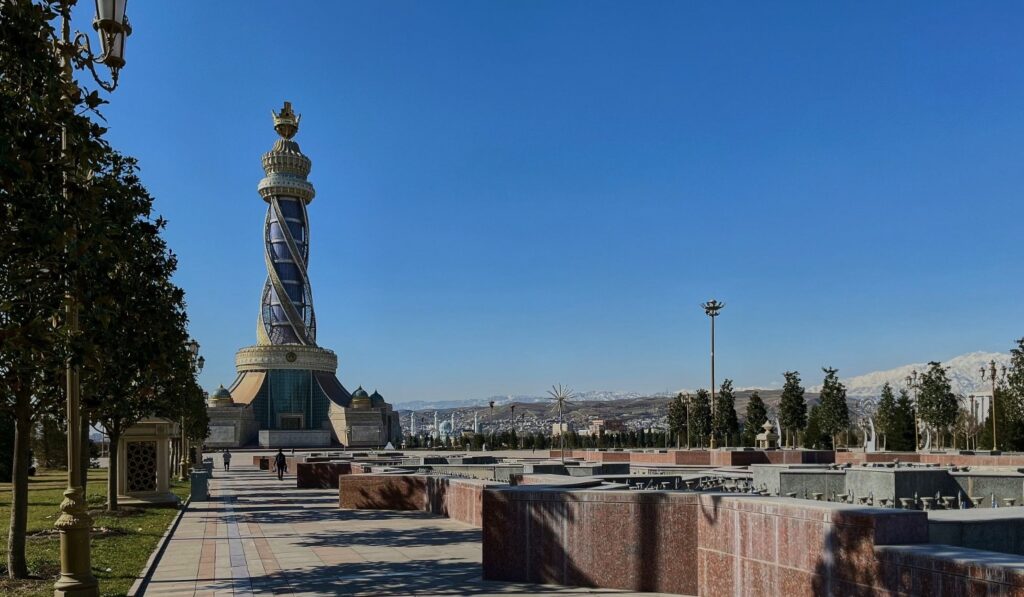 A tall, ornate stone monument with a golden crown on top stands in a modern public plaza in Dushanbe, Tajikistan, with snow-capped mountains in the background.