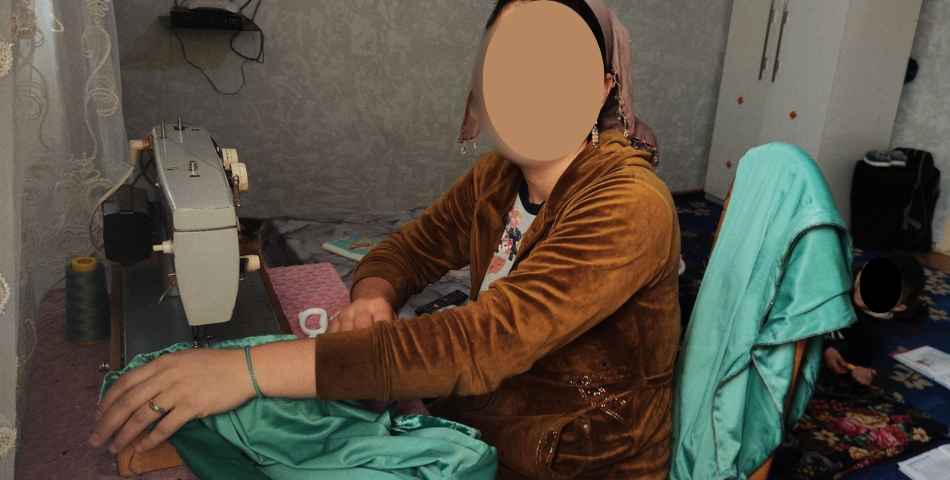 A woman in a brown velvet jacket and headscarf sitting at a vintage sewing machine, working on green fabric in her home workshop with her face blurred for privacy.