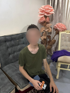A young man in a green t-shirt sitting on a gray couch in a room decorated with large decorative pink roses, with his face blurred for privacy.
