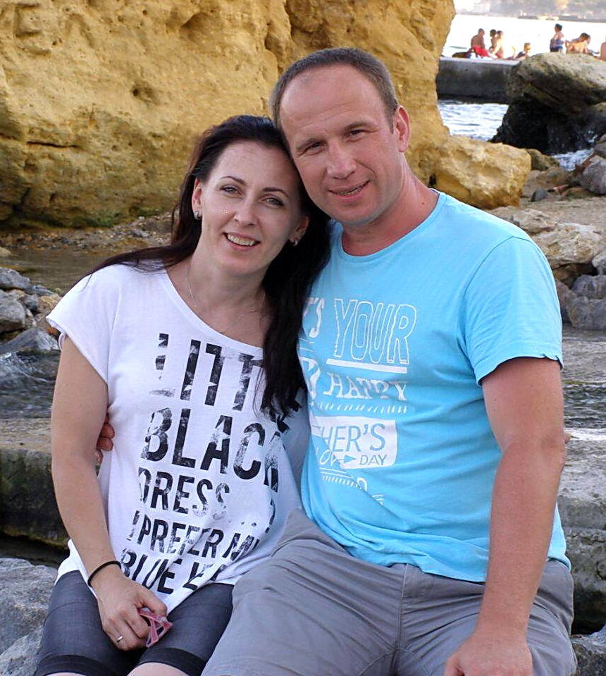 A smiling middle-aged man, Dmytro Panychuk, in a light blue t-shirt sitting closely with a smiling dark-haired woman in a white graphic tee in front of rocky cliffs.