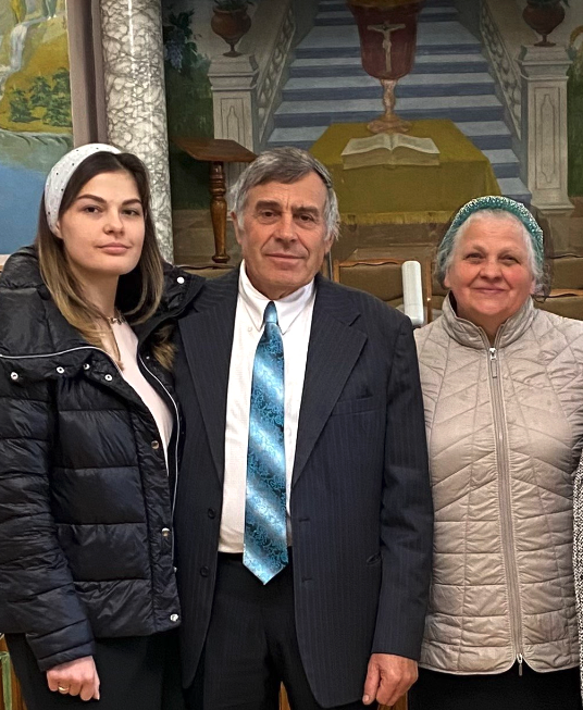 Pastor Leonid Skumatov stands with his wife Liubov and daughter Daria inside their church building, with religious artwork and an altar visible in the background.