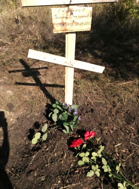 A simple wooden cross marking the grave of Pastor Leonid Skumatov in a yard in Myrnohrad, Ukraine, decorated with fresh red roses and small plants.