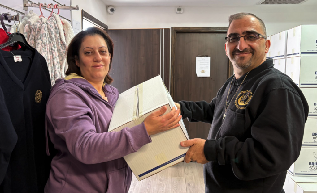 A smiling ministry leader wearing a black hoodie with a cross necklace hands a large, white cardboard food assistance package to a woman in a purple jacket inside a room in Israel.
