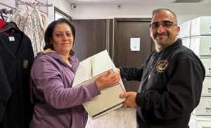 A smiling ministry leader wearing a black hoodie with a cross necklace hands a large, white cardboard food assistance package to a woman in a purple jacket inside a room in Israel.