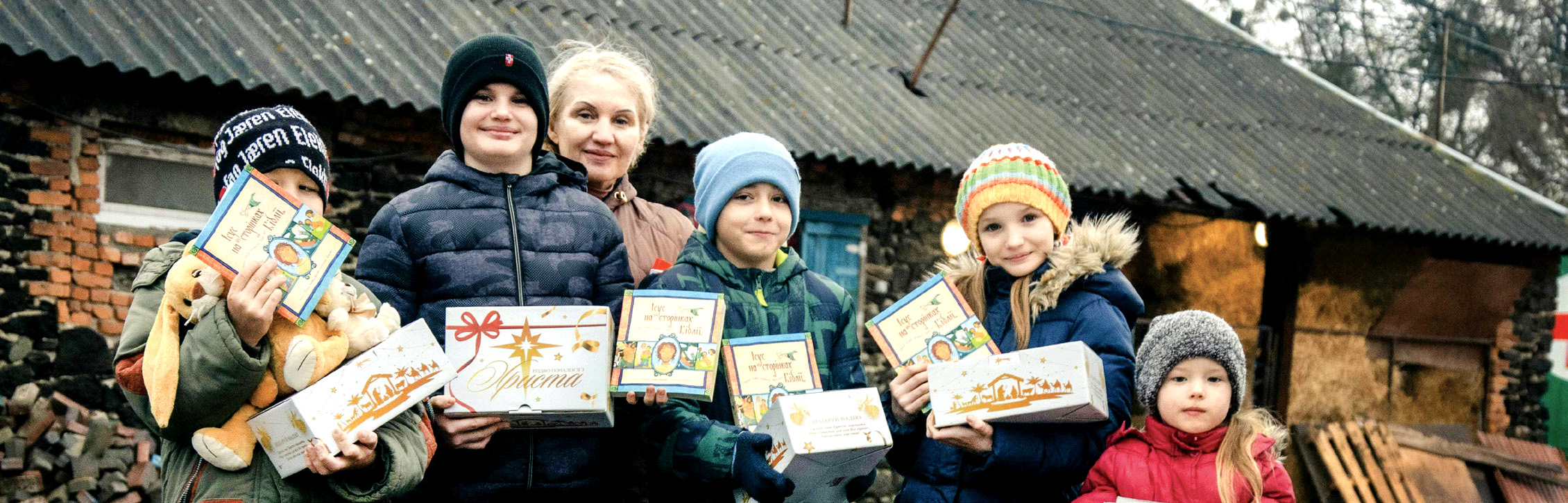 A group of smiling Ukrainian children standing outside together, happily holding and showing Christmas gift boxes during a Mission Eurasia relief distribution.