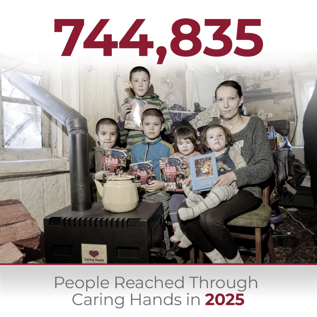 A family gathered next to a small wood-burning stove inside a rustic home, with a mother sitting on a chair holding a young child while three other children stand beside them holding up identical children's Bibles, with text overlay reading 744,835 People Reached Through Caring Hands in 2025.