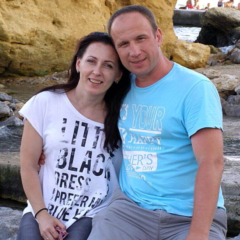 A smiling middle-aged man, Dmytro Panychuk, in a light blue t-shirt sitting closely with a smiling dark-haired woman in a white graphic tee in front of rocky cliffs.