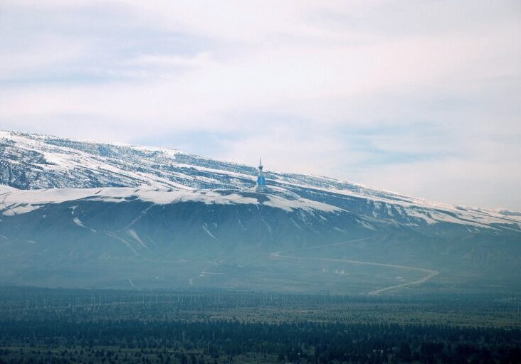 A wide scenic view of the snow-capped Kopet Dag mountains under a hazy sky, overlooking a green valley with a winding road and a distant monument.