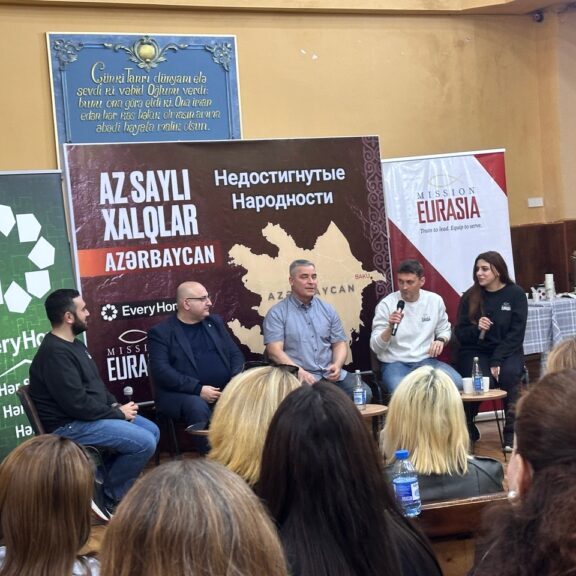 A panel of ministry leaders speaking at a Mission Eurasia event in Azerbaijan, with banners displaying maps of unreached people groups and ministry branding in the background.
