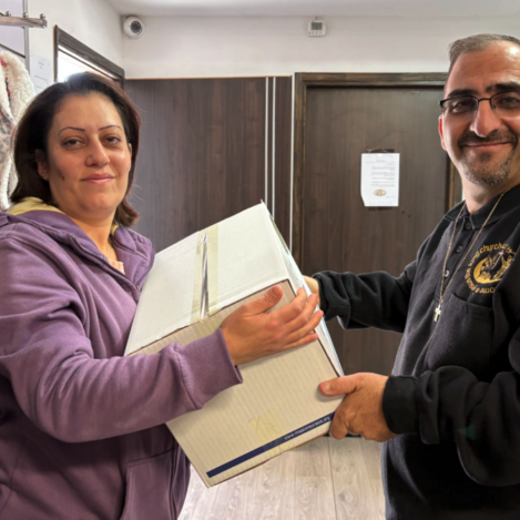 A smiling ministry leader wearing a black hoodie with a cross necklace hands a large, white cardboard food assistance package to a woman in a purple jacket inside a room in Israel.
