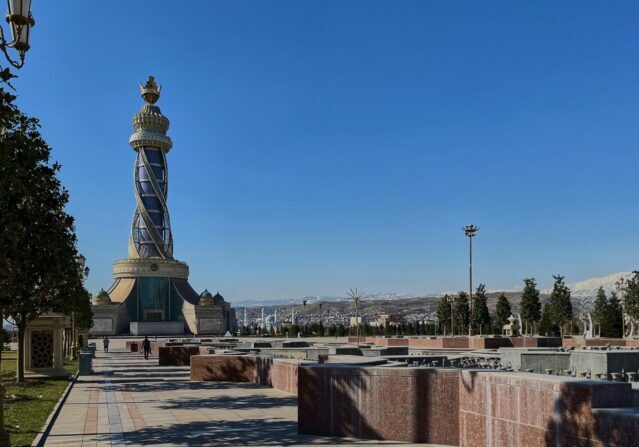 A tall, ornate stone monument with a golden crown on top stands in a modern public plaza in Dushanbe, Tajikistan, with snow-capped mountains in the background.