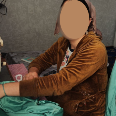 A woman in a brown velvet jacket and headscarf sitting at a vintage sewing machine, working on green fabric in her home workshop with her face blurred for privacy.