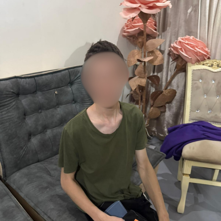 A young man in a green t-shirt sitting on a gray couch in a room decorated with large decorative pink roses, with his face blurred for privacy.