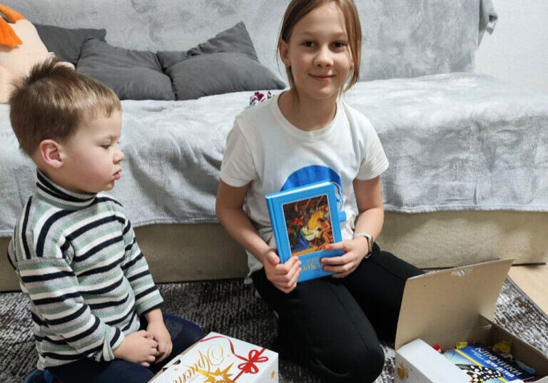 Two young children sitting on the floor indoors, with a girl holding a colorful book and a young boy looking at an open gift box containing various items.